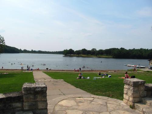 A scenic view of a lake with people swimming and relaxing on the shore, surrounded by green grass and trees.