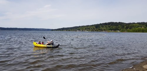 A person and a child paddle in a yellow kayak on a calm lake, surrounded by green hills and a clear sky.