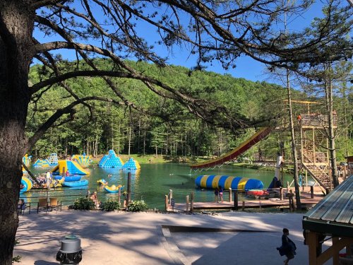 A scenic view of a lake with inflatable water attractions, surrounded by trees and hills on a sunny day.