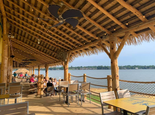 A waterfront dining area with wooden beams, fans, and tables, overlooking a calm lake on a sunny day.