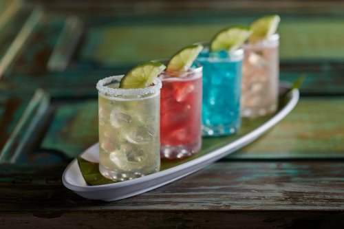Four colorful cocktails with lime slices, served on a white platter against a rustic wooden background.