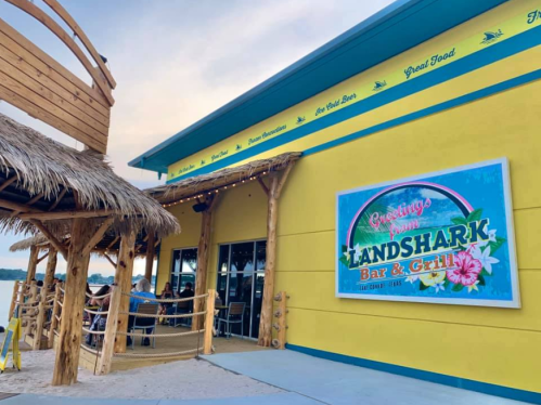 Exterior of Landshark Bar & Grill with a colorful sign, thatched roof, and outdoor seating by the water.