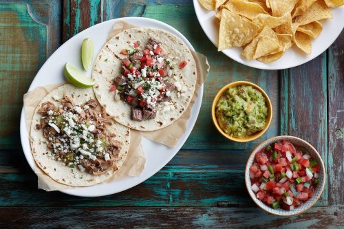 Two tacos filled with meat, topped with cheese and pico de gallo, served with lime, guacamole, and tortilla chips.