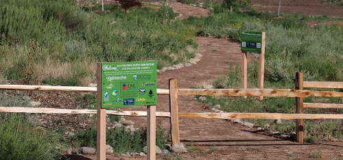 Pathway through a garden with informational signs and wooden fencing, surrounded by greenery and plants.