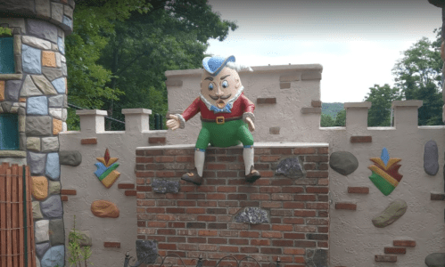 A whimsical character statue in colorful attire sits atop a brick wall, surrounded by a decorative stone fence.
