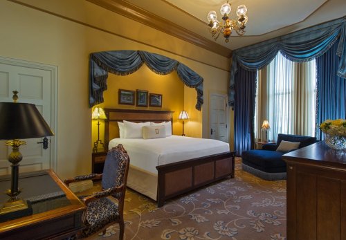 Elegant hotel room featuring a king bed, vintage decor, a seating area, and large windows with blue drapes.