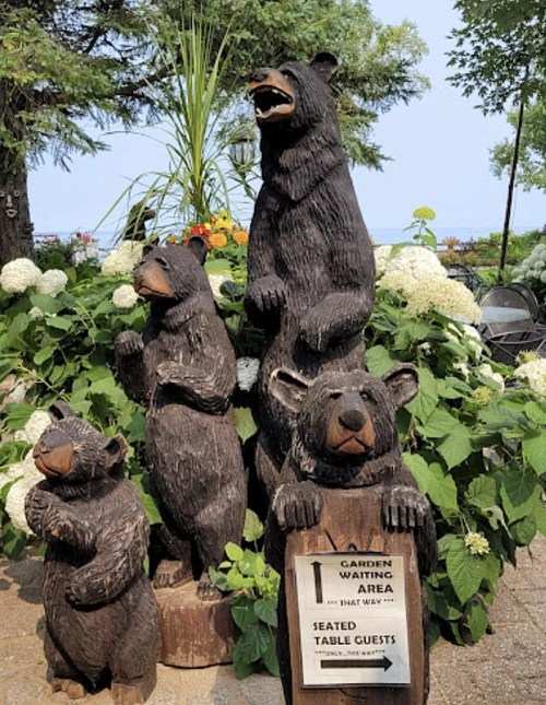 Wooden bear sculptures in a garden, with a sign indicating the waiting area for seated table guests.