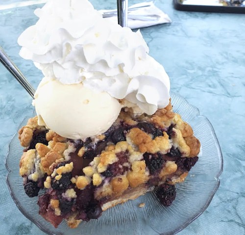 A slice of berry crumble topped with a scoop of vanilla ice cream and whipped cream, served on a glass plate.