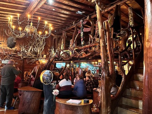 Cozy rustic restaurant interior with wooden decor, chandeliers, and patrons seated at large wooden tables.