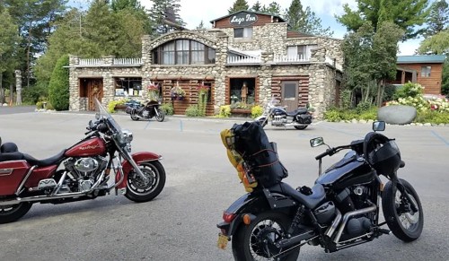Two motorcycles parked in front of a rustic stone inn surrounded by trees and flowers.