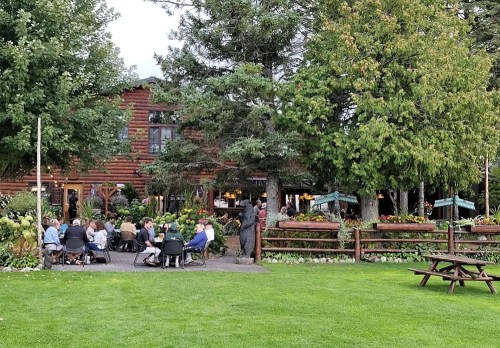 A cozy outdoor gathering at a rustic lodge, surrounded by greenery and flowers, with people seated at tables.