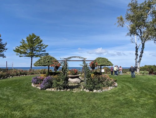 A landscaped garden with flower beds, two canopied seating areas, and people enjoying the view by the water.