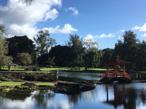 A serene park scene featuring a red gazebo by a pond, surrounded by lush greenery and a stone bridge in the background.