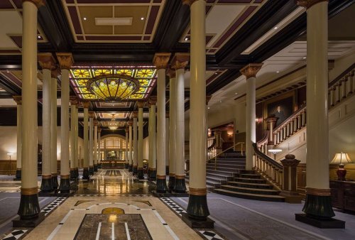 Elegant hotel lobby featuring tall columns, a stained glass ceiling, and a grand staircase. Warm lighting enhances the ambiance.