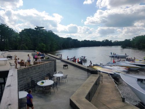 A riverside scene with people socializing, boats docked, and a sunny sky with scattered clouds.