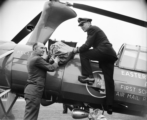 Two men loading mail bags into a helicopter, with one man seated on the helicopter's edge and the other standing beside it.