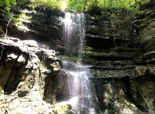 A serene waterfall cascading down rocky cliffs, surrounded by lush green foliage and dappled sunlight.