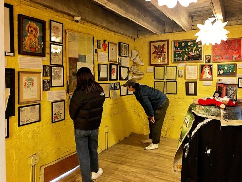 Two people examine a colorful art gallery wall filled with framed artworks and information on a yellow background.