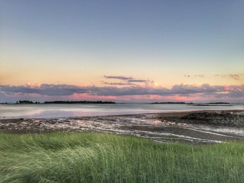 A serene coastal scene at sunset, featuring calm waters, distant islands, and grassy foreground under a colorful sky.
