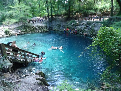 A natural spring with clear blue water, surrounded by lush greenery and people swimming and relaxing.