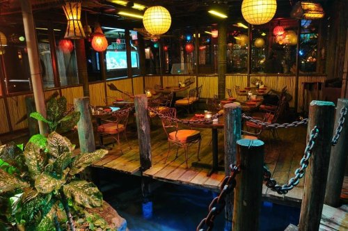 Cozy restaurant interior with wooden decor, colorful lanterns, and tables surrounded by plants and water features.
