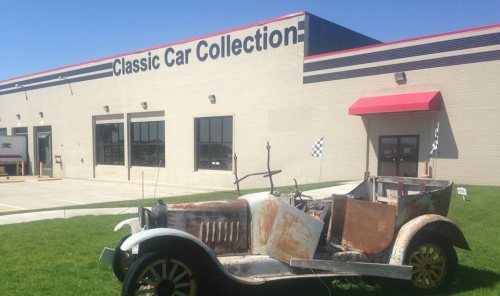 An old, rusty car in front of a building labeled "Classic Car Collection" on a sunny day.