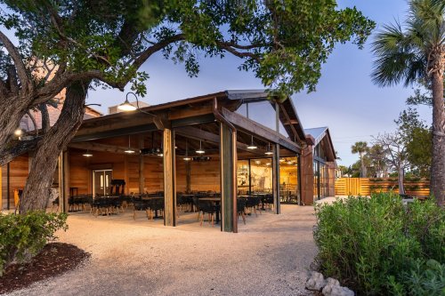 A modern restaurant with wooden architecture, outdoor seating, and lush greenery, set against a clear sky.