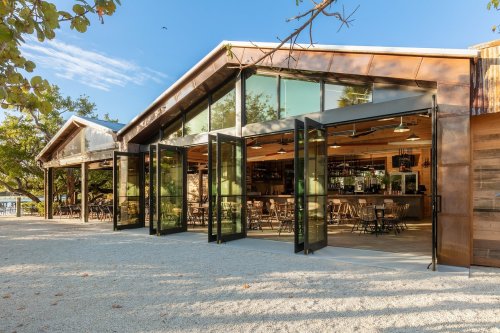 Modern restaurant with large glass doors open to a patio, surrounded by trees and a clear blue sky.