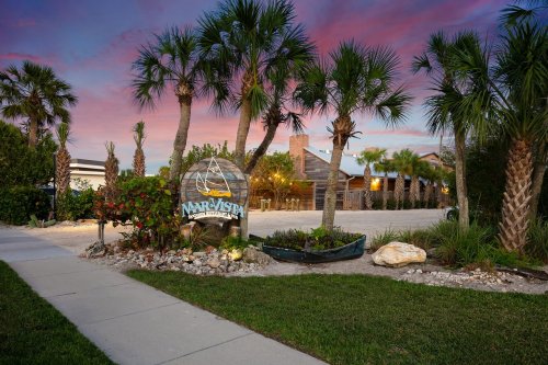 A welcoming sign for a coastal venue surrounded by palm trees and colorful flowers, with a sunset sky in the background.