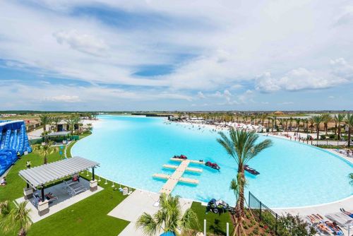 A large, blue lagoon with palm trees, lounge chairs, and a water slide under a bright sky.
