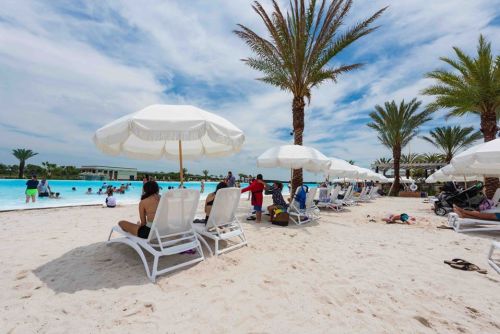 A beach scene with lounge chairs, palm trees, and people enjoying a lagoon under a partly cloudy sky.