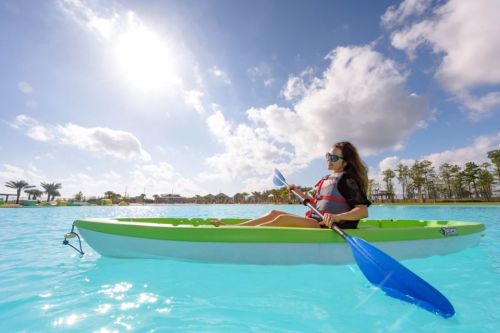 A woman in a kayak paddles on a bright blue lake under a sunny sky with fluffy clouds.