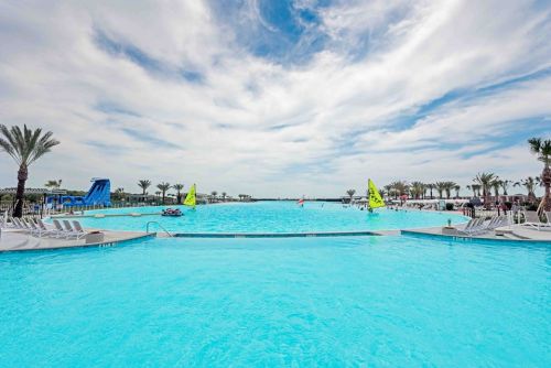 A large, bright blue pool surrounded by palm trees and lounge chairs, with sailboats in the distance under a cloudy sky.