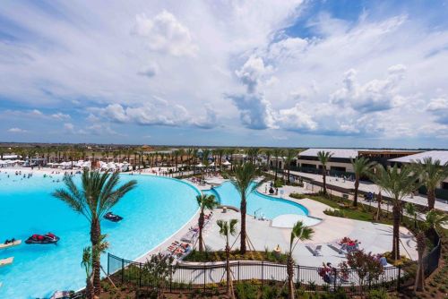 A large resort pool surrounded by palm trees, with people enjoying the water and a cloudy sky in the background.
