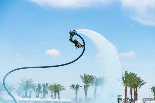 A person performs a stunt on a jetpack, soaring above water with palm trees in the background under a blue sky.