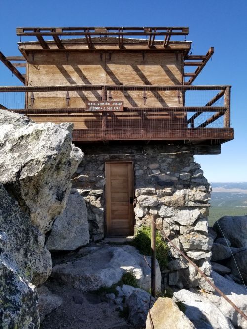 A wooden lookout tower atop rocky terrain, with a door and scenic views in the background under a clear blue sky.