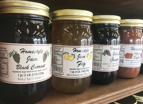 Four jars of homemade jams and jellies on a shelf, labeled black currant, fig, grape, and apple butter.