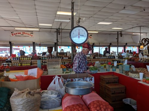 A farmer weighs produce at a vibrant market filled with fruits, vegetables, and jars of preserves.