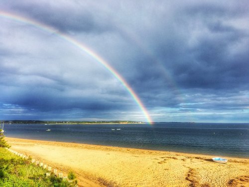 A beach scene with a rainbow arching over the water, under a cloudy sky. Boats are visible in the distance.
