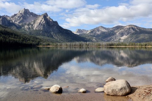 A serene lake surrounded by mountains, with clear reflections and a rocky shore under a partly cloudy sky.