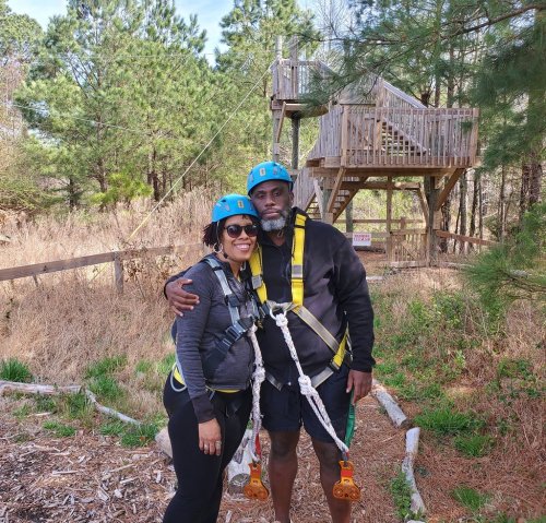 A couple in harnesses and helmets smiles together in a forested area near a zipline platform.