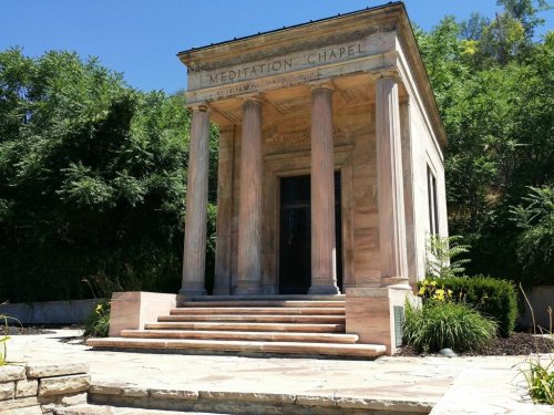 A stone meditation chapel with columns, surrounded by greenery and a stone pathway leading to the entrance.