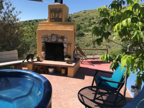 A cozy outdoor space featuring a hot tub, stone fireplace, and seating area with a scenic green hillside in the background.