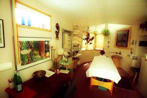 A cozy treatment room with a massage table, artwork on the walls, and natural light streaming in through a window.