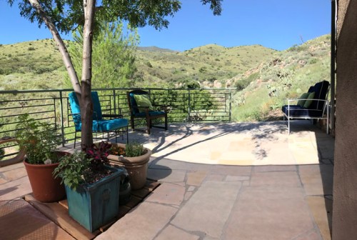 A serene outdoor patio with potted plants, chairs, and a view of green hills under a clear blue sky.