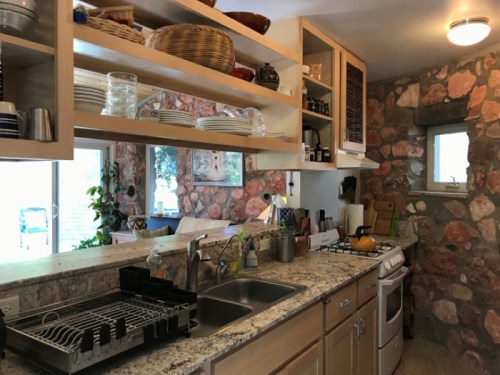 A cozy kitchen with stone walls, open shelves, and a sink, featuring dishes and cooking utensils. Natural light from a window.