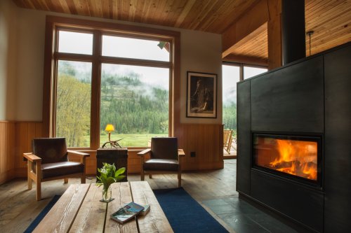Cozy living room with a fireplace, wooden decor, and large windows showcasing a misty forest view.