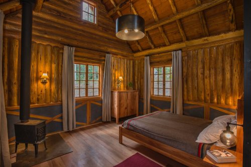 Cozy wooden cabin interior with a bed, windows, and a small stove, featuring warm lighting and rustic decor.