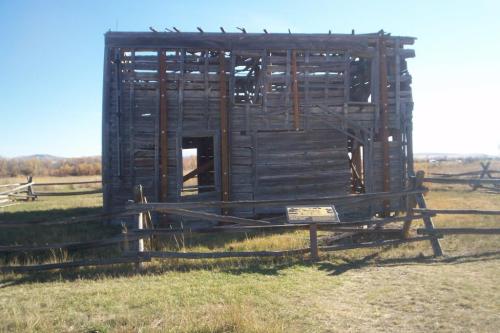 A weathered, partially collapsed wooden barn surrounded by a fence and grassy field under a clear blue sky.