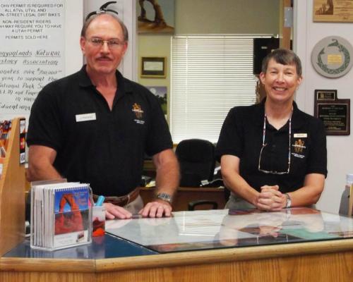 Two smiling staff members stand behind a reception desk, wearing matching black shirts with name tags.